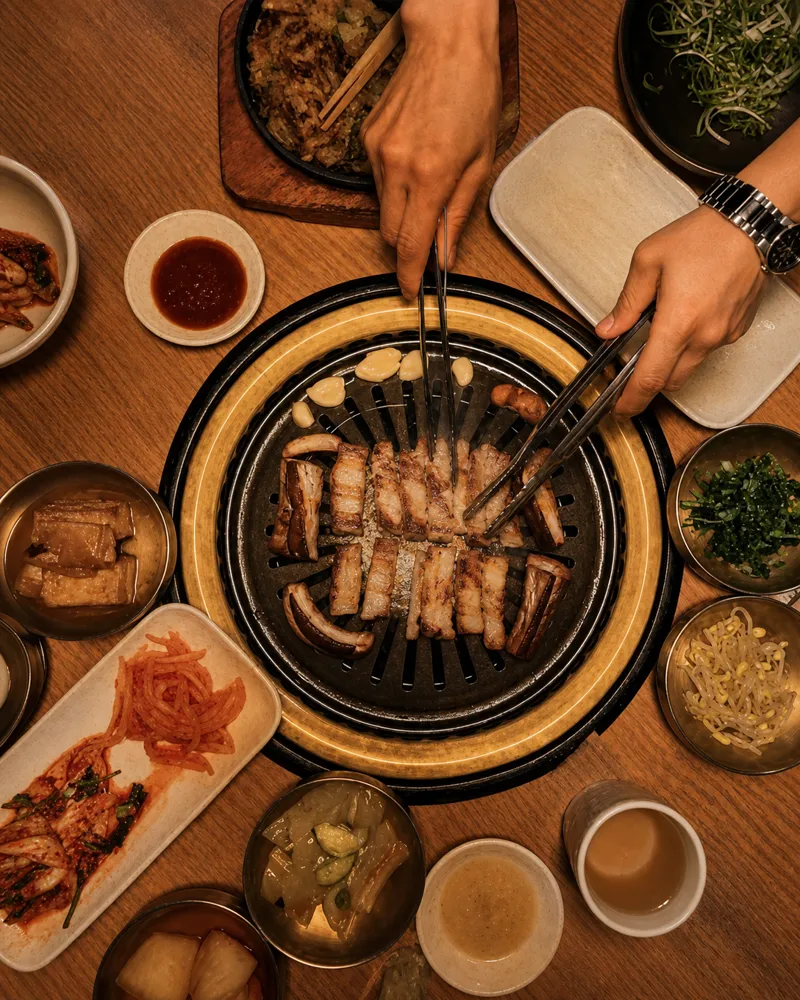 Pork belly grilling on the tabletop smokeless grill, tongs turning the meat, banchan all around
